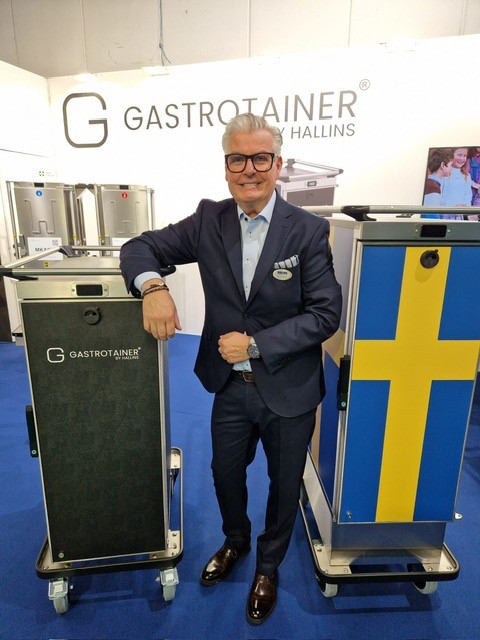 Man i kostym står vid två serveringsvagnar med varumärket Gastrotainer, en prydd med svensk flagga.
