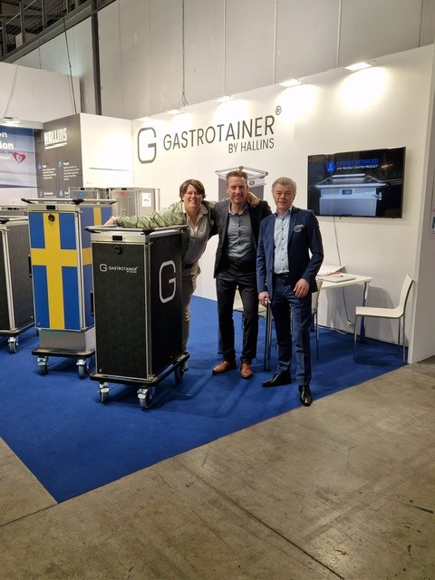 Tre personer poserar vid serveringsvagnar i Hallins Gastrotainer-monter på mässa.