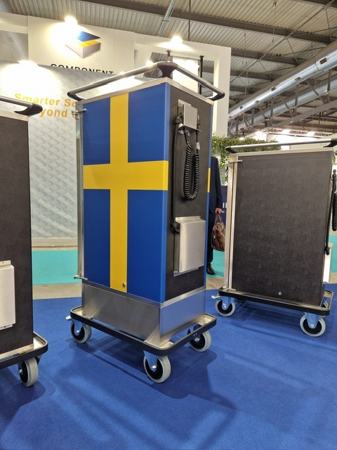 Serveringsvagn med svensk flagga på sidan i mässmonter med blå matta och industrimiljö.